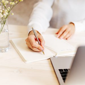 Ein Schreibtisch aus hellem Holz, Frauenhände, die von inks ins Bild hineinragen, schreiben einen Brief. Die Frau trägt helle Kleidung. Rechts steht eine Vase mit Blumen.